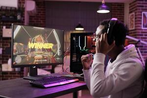 africano americano jugador emocionado acerca de victorioso en línea multijugador partido en contra otro jugadores hombre viendo ganador mensaje en juego de azar ordenador personal computadora monitor en neón luces vivo habitación foto