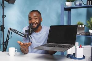 Smiling social media influencer promoting laptop while recording for social media. African american streamer advertising portable computer while live broadcasting and looking at camera photo