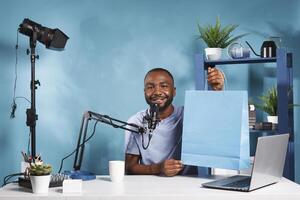 Smiling blogger holding shopping bag while recording unpacking for internet channel. Influencer streaming giveaway contest, showing package and looking at camera while streaming live photo