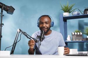 africano americano blogger en auriculares conectando con suscriptores en línea y mirando a cámara. flámula vistiendo auriculares Hablando en micrófono y Bebiendo café mientras rodaje foto