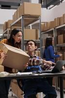 Warehouse asian workers holding parcel and scheduling delivery on laptop. Storehouse employees discussing orders checklist and analyzing pick ticket on computer in storage room photo