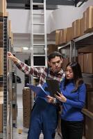 Warehouse employees managing inventory tracking with goods checklist. Storehouse asian man and woman workers picking order while standing in storage room aisle with shelves full of cardboard boxes photo