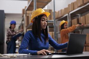 Asian retail storehouse employee analyzing orders checklist on laptop while working in storage room. Warehouse manager supervising inventory management while using computer photo