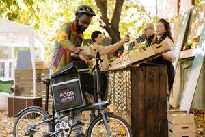negro masculino mensajero poniendo caja de orgánico productos en mochila para entregando orden a clientela, utilizando bicicleta. joven alegre repartidor trabajando en agricultores mercado verduras envío. foto