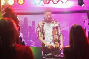 Dj on stage controlling music while crowd partying and dancing in nightclub. Man musician mixing sound on console while performing at live concert in club with spotlights photo