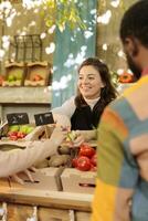 Smiling woman vendor offering customer to try out small piece of organic apple while selling fresh natural fruits and vegetables at harvest fair festival, selective focus. Tasting during shopping. photo