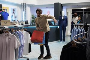 Happy young caucasian man dancing with paper packages in shopping center after buying fashionable apparel. Satisfied joyful customer carrying bags with purchased trendy outfit photo