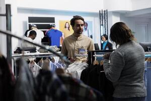 ropa Tienda consultor estrella de guía cliente mediante mercancías y utilizando digital tableta. africano americano mujer hojeada vestir estante y hablando con empleado mientras compras en centro comercial foto