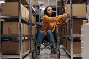 mujer silla de ruedas usuario tomando cartulina caja desde estante a preparar cliente orden para despacho en depósito. asiático paquete manipulador participación parcela, trabajando en invalidez simpático almacén foto