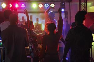 clubbers ir de fiesta y bailando mientras asistiendo DJ actuación en Club nocturno. joven personas multitud en pie con elevado manos en pista de baile iluminado con focos a discoteca foto