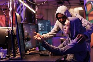 Two hackers collaborating while breaching data and coding malicious software on computer. Diverse criminals working together while programming ransomware in abandoned warehouse photo