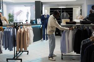 africano americano hombre buscando camisa Talla mientras elegir formal vestir atuendo en ropa almacenar. boutique cliente hojeada estante con colgando vestir en compras centro comercial toma de corriente foto
