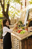 Mature man and young woman wearing apron and arranging greenmarket stall with eco friendly bio food products. Female vendor holding bunch of carrots, preparing farmers market booth for customers. photo