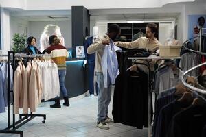 Clothing store customer trying on shirt and getting advice from boutique employee. African american man holding apparel to body while choosing size with fashion outlet assistant photo