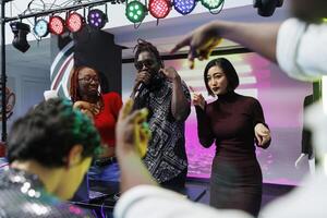 African american musician singing in microphone at electronic music festival live concert in nightclub. Diverse man and women partying and performing on stage at event in club photo