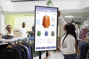 Female customer reviewing clothes to purchase from self ordering kiosk service, looking at trendy fashion items on touch screen board. Young client checking products on monitor. photo