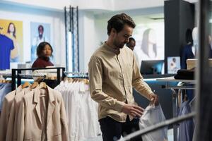 hombre seleccionando de moda atuendo en ropa Tienda y hojeada estante con camisas joven cliente examinando vestir en perchas mientras elegir formal vestir en compras centro comercial Departamento foto