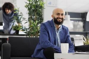 Smiling arab business entrepreneur looking at camera while sitting on couch in office. Start up company successful executive manager wearing suit portrait in coworking space photo