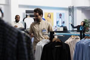 Clothing store manager using digital tablet and organizing merchandise to attract customers. Shopping center employee managing inventory and examining apparel hanging on rack photo