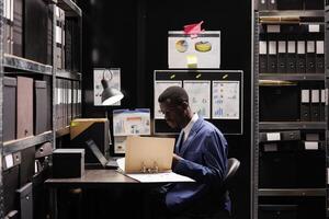 African american manager checking administrative files, analyzing bureaucracy record late at night in arhive room. Corporate worker in formal suit organizing accountancy documents. Depository concept photo