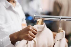 africano americano mujer asistente ajustando chaquetas en perchas en compras centro comercial. ropa Tienda empleado colgando de moda chaquetas americanas desde nuevo colección para rebaja en monitor estante cerca arriba foto