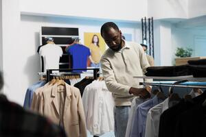 africano americano hombre elegir formal atuendo y hojeada camisas colgando en estante. joven elegante comprador buscando para de moda prenda mientras compras en Al por menor centrar Departamento Tienda foto