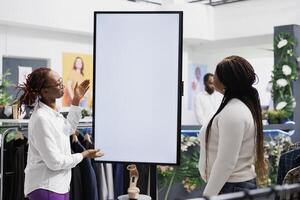 Assistant helping customer choosing shoes, showing style and size options on smart white empty display. Clothing store employee showcasing promotion on blank whiteboard screen mock up photo