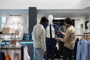 ropa Tienda asistente exhibiendo cliente elegante traje en maniquí. compras centrar ropa de caballero boutique diverso empleado y cliente examinando formal chaqueta calidad mientras elegir atuendo foto