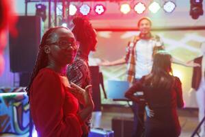 africano americano mujer en pie en pista de baile demostración con mano a sí misma en Club nocturno. joven discotequero teniendo divertido, relajante y ir de fiesta en club con focos, disfrutando la vida nocturna foto