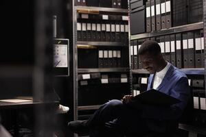 africano americano gerente organizando corporativo archivos, analizando burocracia grabar en archivar habitación. empresario vistiendo formal traje comprobación contabilidad documentos en oficina depositario foto