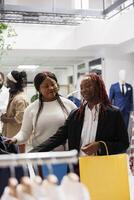 africano americano mujer compras para ropa y chateando en centro comercial. novias clientes buscando para vestir colgando en estante y elegir atuendo juntos en Moda boutique foto