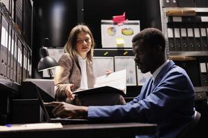 Police officers analyzing criminology report, working overtime at criminal case in arhive room. Diverse private investigators discussing crime scene, checking police evidence. Law enforcement concept photo