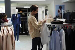 Clothing store customer holding formal shirt on hanger and looking at size label. Young stylish man checking apparel and choosing fashionable garment in shopping mall department photo