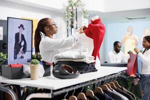 africano americano mujer vendaje maniquí en rojo pull-over en ropa almacenar. compras centro comercial asistente poniendo hembra casual blusa en tonto modelo a monitor vestir para rebaja foto