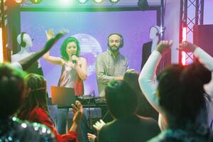 Young man and woman band performing on stage in nightclub at techno music festival. People partying on dancefloor while enjoying singer and dj playing at live concert in club photo