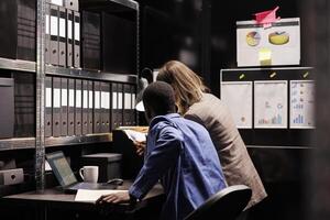 Depository employees working overhours at bureaucracy record, analyzing administrative report in storage room. Diverse bookkeepers checking accountancy files, organizing corporate documents photo