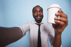 negro hombre teniendo un llamada mientras participación arriba taza de café en contra aislado antecedentes. sonriente africano americano vestido en blanco camisa haciendo un selfie foto con bebida envase en su mano.