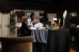 Hip level shot of cozy tailoring studio with blurry african american dressmaker in the background studying sartorial designs. Couturer inspecting sketches of upcoming fashion collection photo