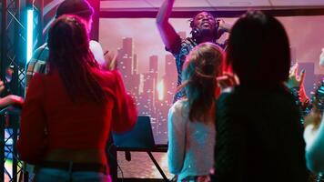 Young man clubbing with people at bar, having fun with DJ mixing station on nightclub stage with disco ball. Funky person creating live performance on audio panel, spotlights at club. photo