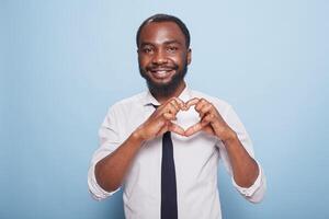 Smiling african american influencer making heart shape fingers to show care for supporting fans. Joyful male blogger in white shirt and black tie doing the hand gesture for love on his chest. photo