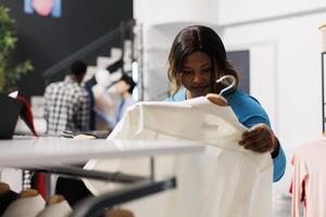 African american customer holding hanger with white shirt, checking items fabric in modern boutique. Shopaholic woman looking at new fashion collection, shopping for casual wear in clothing store photo