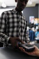 African american customer typing credit card details on pos to pay for stylish clothes, making contactless payment at store counter. Shopaholic man buying fashionable items for casual wear in boutique photo