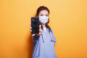 Nurse wearing scrubs and face mask, gripping cell phone with blank screen. Medical assistant stands in front of isolated background, looking at camera and displaying empty mobile device screen. photo
