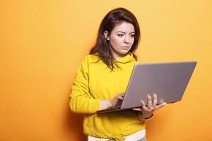 Female freelancer utilizing modern technology by using digital laptop. Portrait of focused woman working online on wireless computer while standing over isolated orange background. photo