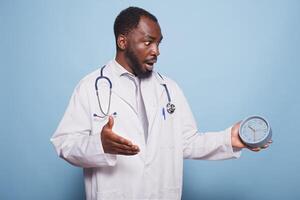 africano americano general facultativo espantosamente mirando a reloj antes de un paciente médico consulta. negro médico aparece a ser en un pánico, corriendo y experimentando un cuidado de la salud emergencia. foto