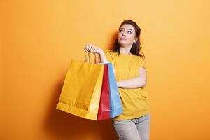 Happy, stylish brunette shopper carries shopping bags and smiles on isolated orange background. Caucasian woman looking attractive and cheerful while grasping colorful parcels. photo