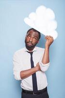 retrato de masculino empresario en blanco camisa mirando arriba a blanco papel idea nube lluvia de ideas mientras en pie en frente de azul antecedentes. africano americano hombre pensando debajo pensamiento burbuja. foto