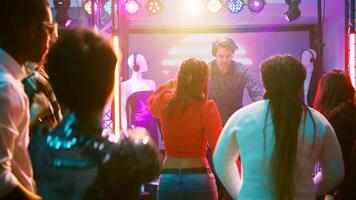 Funky DJ jumping on stage mixing beats, having fun with crowd of people at nightclub. Young men and women partying together and dancing on electronic sounds, entertainment. Handheld shot. photo