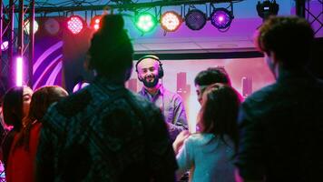 Male DJ putting music at club party, mixing electronic sounds at audio equipment station. Diverse group of friends jumping around and dancing on dance floor, clubbing entertainment. photo