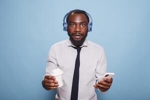 Shocked black man in white shirt looking at camera while holding cell phone for streaming music to wireless headphones. Young freelancer with a takeaway cup, having coffee and browsing internet. photo